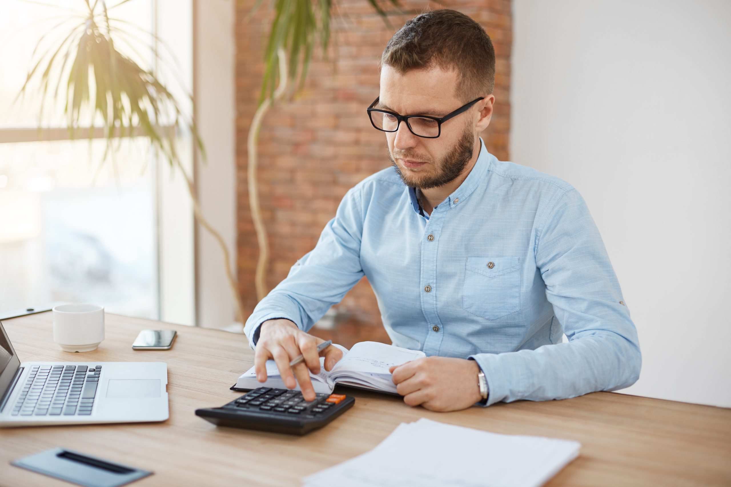 Adult serious bearded caucasian finance manager in glasses and blue shirt sitting in light comfortable company office, cheking month profits with calculator, writing down results in notebook, speding productive morning